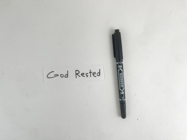 A small strip of white paper with the handwritten text "God Rested" next to a black marker.