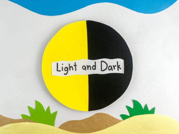 A finished paper craft of a circle split into yellow and black halves with a label that says "Light and Dark," set against a background of paper hills, grass, and sky.