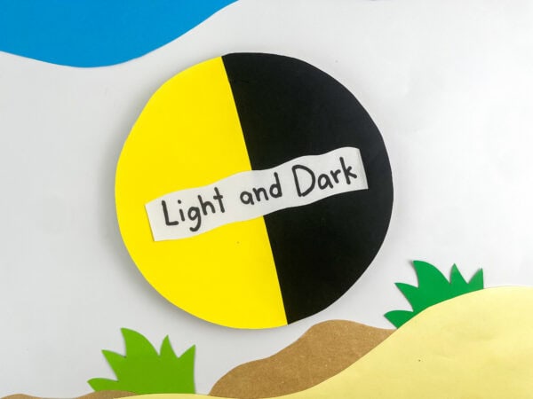 A slightly angled shot of the finished "Light and Dark" paper circle craft over a decorative background of paper hills and blue sky.