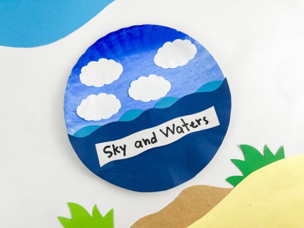 A completed "Sky and Waters" paper plate craft displayed against a decorative backdrop featuring paper cutouts of green grass and sandy shores.