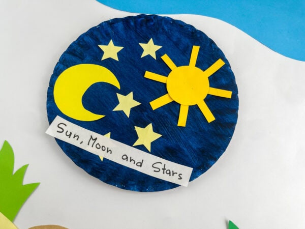 An angled shot of the completed "Sun, Moon and Stars" paper plate craft on a white surface with paper grass cut-outs.