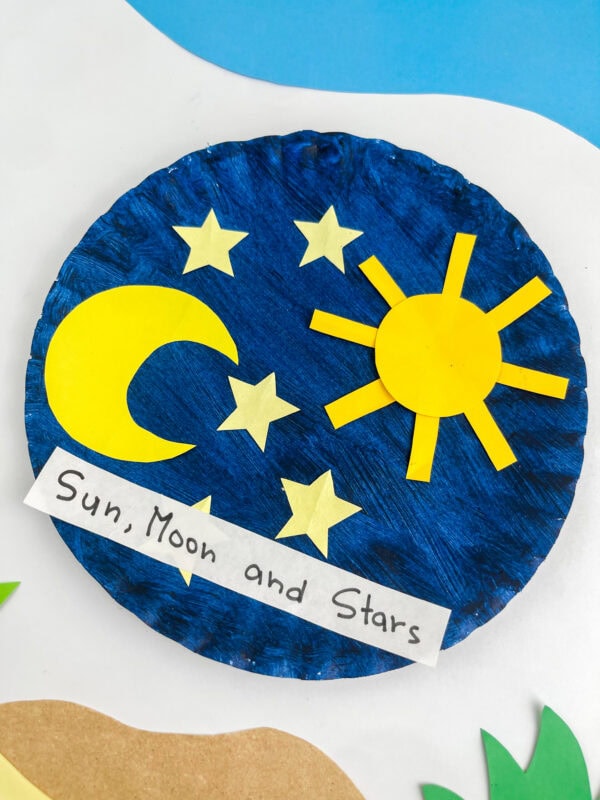 A close-up, angled view of the yellow paper sun, moon, and stars craft on a painted blue paper plate.
