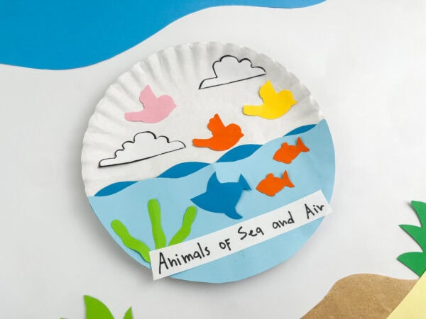 An angled close-up shot of the paper plate craft with decorative blue and brown paper background cutouts simulating sky and shore.