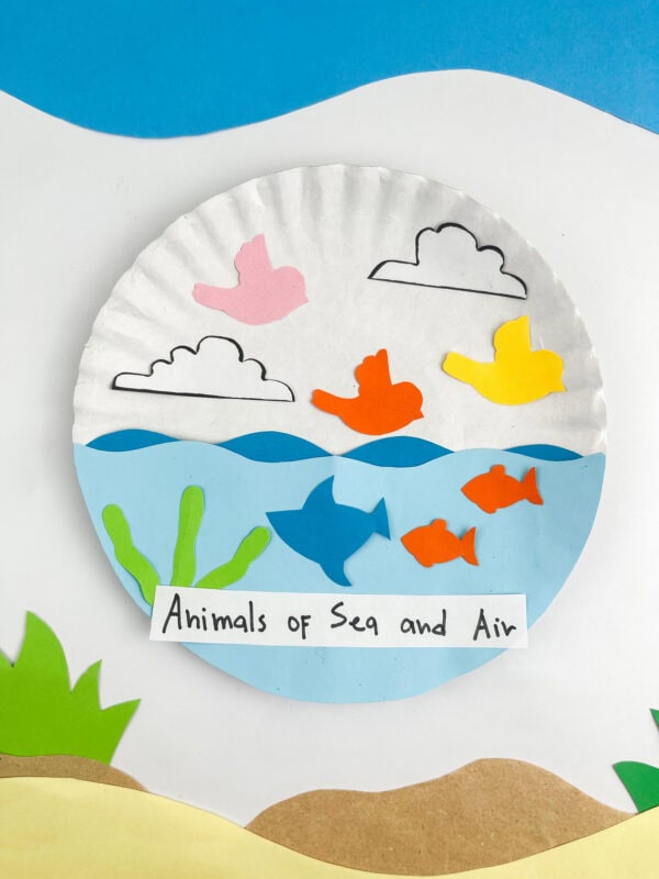 The finished paper plate craft centered flat against a themed backdrop of blue water and a sandy shoreline.