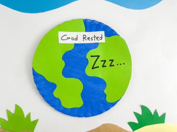 A finished paper plate craft representing Earth, with paper cutouts of grass and a sky backdrop.