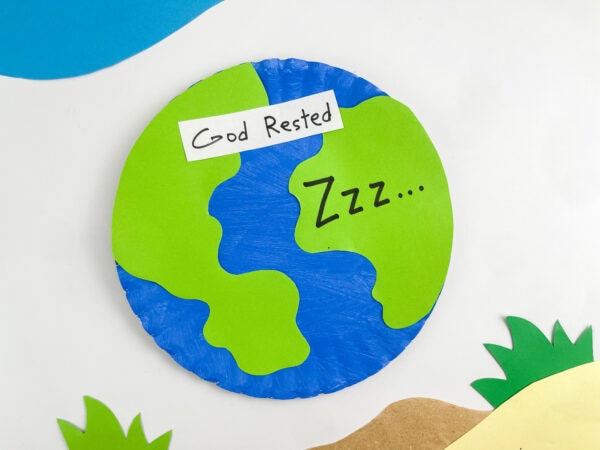 A completed paper plate Earth craft featuring a "God Rested" label and "Zzz..." text, framed by paper grass and sky cutouts.