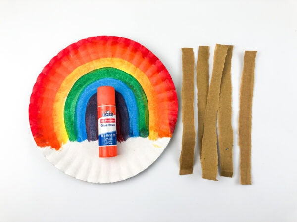 Rainbow-painted paper plate, a glue stick, and cardboard strips arranged side by side.
