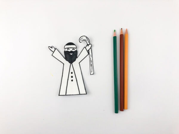 A black-and-white paper cutout of a robed man with a headband, beard, and staff, beside green and orange pencils.