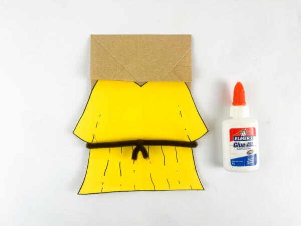 assembling Deborah paper bag puppet body with yellow robe and pipe cleaner belt on kraft paper bag
