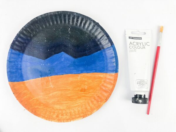 Paper plate painted with blue, black, and orange sections with paintbrush.