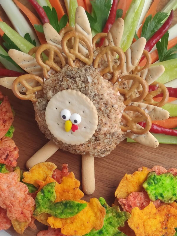 Turkey cheese ball appetizer decorated with a cracker face, pretzels, fresh vegetables, and colorful leaf-shaped chips.