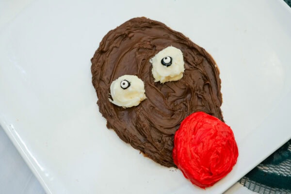 Close-up of a chocolate frosting reindeer face with white icing patches, candy eyes, and a bright red nose.
