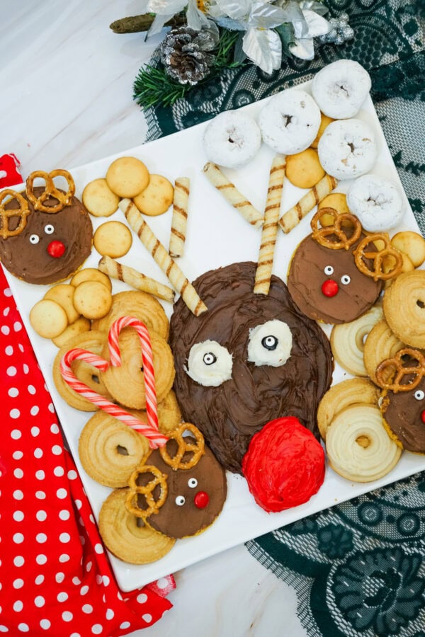 Close-up of Rudolph's chocolate frosting face surrounded by rolled wafer antlers, decorated reindeer cookies, powdered doughnuts, and holiday snacks.