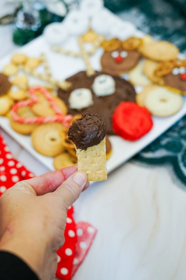 Hand holding a rectangular cracker topped with chocolate frosting in front of a Christmas reindeer dessert platter.